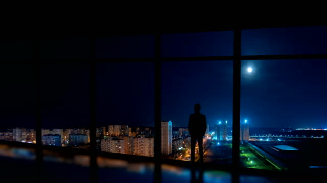 The-man-standing-near-the-window-on-the-night-city-background.-time-lapse