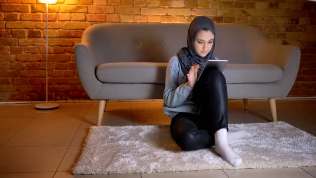 Closeup-shoot-of-young-attractive-muslim-female-in-hijab-smiling-and-scrolling-on-the-phone-while-sitting-on-the-floor-indoors-at-cozy-home