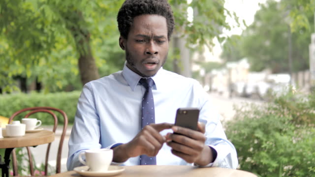 Hombre-de-negocios-africano-molesto-por-la-pérdida-en-el-teléfono-inteligente,-sentado-en-al-aire-libre-Cafe