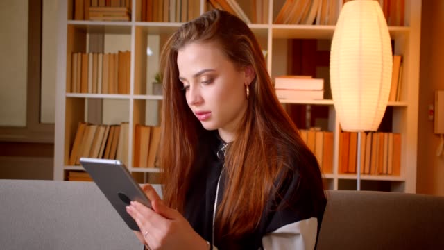 Portrait-of-young-teenage-girl-working-with-tablet-sitting-on-sofa-on-bookshelves-background-watches-into-camera-at-home.