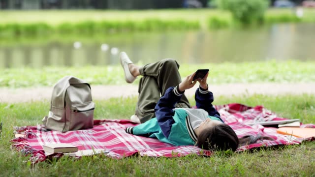 Entspannte-asiatische-College-Mädchen-ruht-auf-Rasen-in-der-Nähe-von-Teich-nach-dem-Unterricht.-Studentin-liegt-auf-karierter-Decke-und-lacht-beim-SMS-Schreiben-auf-dem-Smartphone