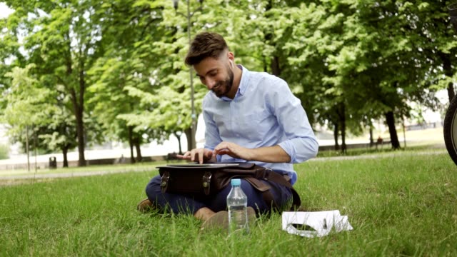 Un-joven-empresario-sonriente-sentado-en-el-parque-durante-el-almuerzo-y-escribiendo-en-la-tableta