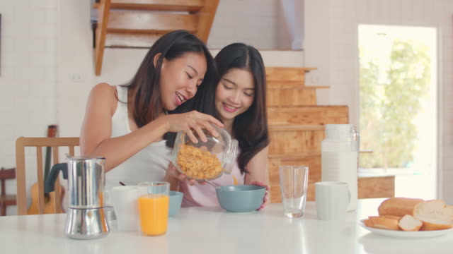 Asian-Lesbian-couple-has-breakfast-drink-juice,-cornflakes-cereal-and-milk-in-bowl-on-table-in-kitchen-in-the-morning.