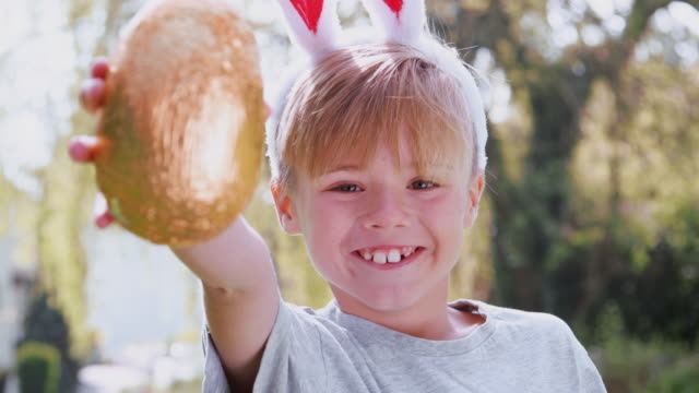 Portrait-of-boy-wearing-bunny-ears-on-Easter-egg-hunt-outdoors-at-home-holding-up-chocolate-egg-to-camera-and-smiling---shot-in-slow-motion