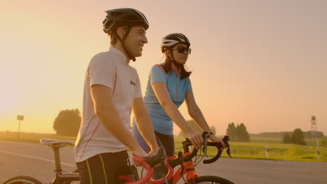 A-man-and-a-woman-in-helmets-with-bicycles-stand-and-talk-at-sunset.-Rest-after-a-bike-ride-on-the-highway.-Track-bikes.-Couple-sports.
