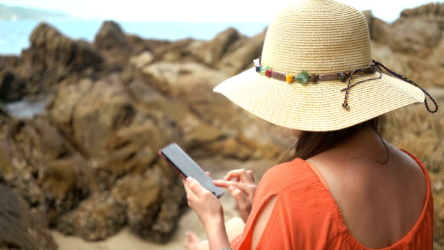 joven-hermosa-mujer-usando-el-teléfono-para-comunicarse-en-las-redes-sociales-y-sentarse-en-una-piedra-cerca-del-mar
