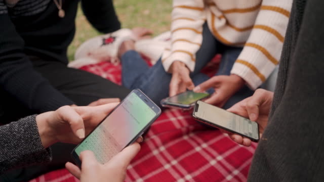 Primer-plano-de-amigos-sentados-juntos-en-una-manta-roja-usando-la-aplicación-móvil-en-el-parque
