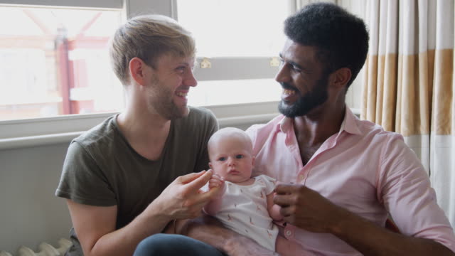 Portrait-Of-Loving-Male-Same-Sex-Couple-Cuddling-Baby-Daughter-On-Sofa-At-Home-Together
