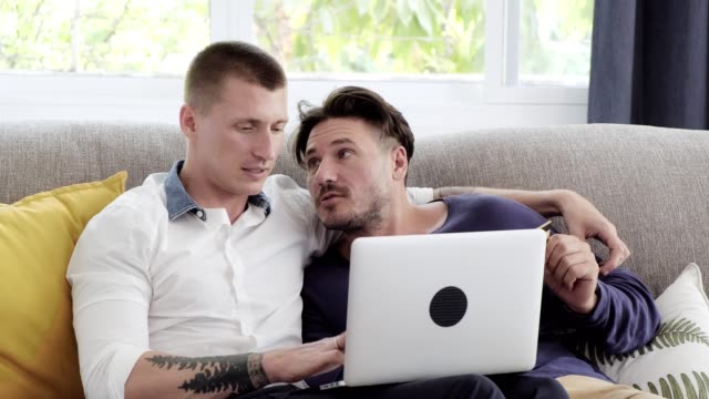 Pareja-gay-relajándose-en-el-sofá-usando-la-computadora-portátil.-Pensar-y-escribir.