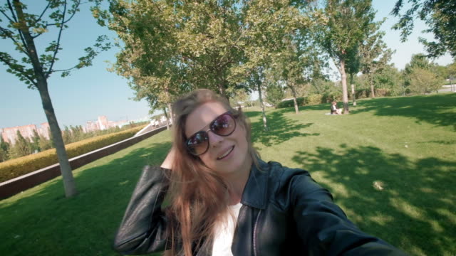 Studentin-im-Park.-Charmantes-Mädchen-in-Lederjacke-spaziert-durch-den-Herbstpark-und-filmt-sich-vor-der-Kamera.-Macht-ein-Selfie-auf-einem-Smartphone-für-soziale-Netzwerke.-zeitlupe.
