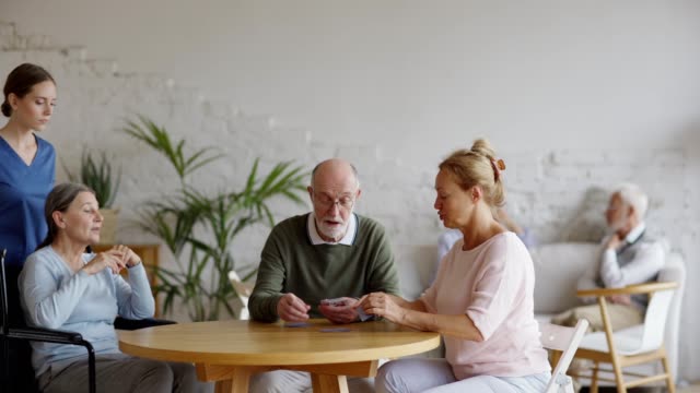 Caregiver-pushing-wheelchair-of-disabled-aged-woman-to-help-her-join-senior-friends-playing-cards-at-table-in-nursing-home.-Elderly-man-and-woman-talking-on-sofa-in-background