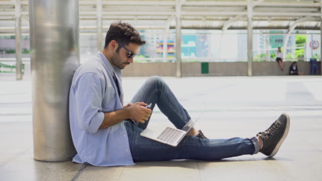 young-businessman-in-smart-casual-wear-sitting-on-street-using-laptop-and-typing-a-message-on-mobile-phone-in-urban-city-outdoors.-freelancer-working-on-floor-outside-Chatting-to-customer.-blue-jeans
