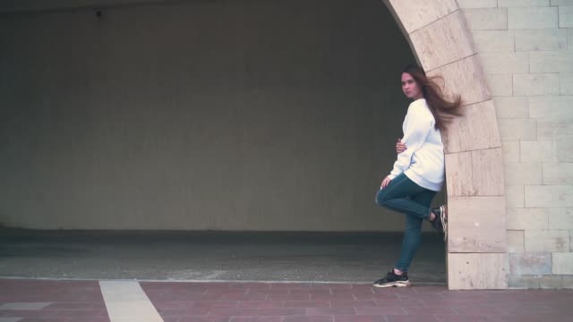 A-girl-with-long-hair-and-a-white-sweatshirt,-standing-with-her-foot-on-the-stone-arch-of-the-building.