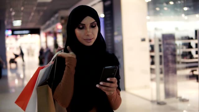 Attraktive-muslimische-Frau-trägt-schwarze-Hijab-Kopftuch-zu-Fuß-in-der-Shopping-Mall-und-mit-dem-Smartphone.-Kommunikation,-Online-Shopping,-Social-Network-Konzept