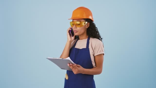 Dark-skinned-woman-industrial-worker-or-builder-in-hard-hat-and-protective-goggles-is-talking-by-smartphone-and-looking-at-papers-on-clipboard
