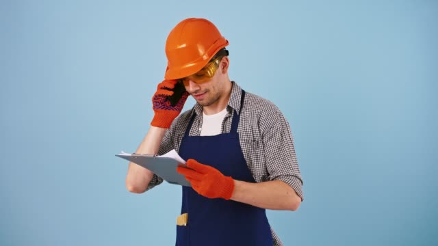 Young-guy-in-safety-wear,-hard-hat,-protective-goggles,-gloves.-Talking-by-mobile-phone,-looking-at-papers-on-clipboard.-Posing-on-blue-background