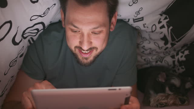 Close-up-of-face-of-a-young-bearded-and-mustashed-man,-using-digital-tablet-under-blanket,-in-his-bed,-at-night.-His-yorkshire-terrier-is-lying-next-to-him