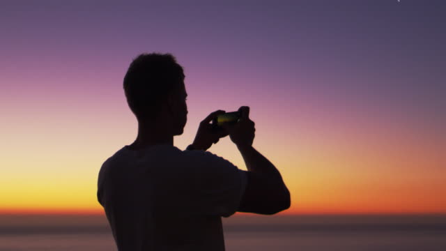 Mann,-der-Panorama-Foto-mit-Handy-am-Strand-bei-Sonnenuntergang