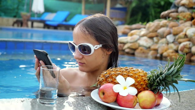 Junge-schöne-Frau-nutzt-Handy-im-Schwimmbad-neben-dem-Glas-des-Wassers-und-der-Platte-mit-tropischen-Früchten-in-Zeitlupe.-HD,-1920-x-1080