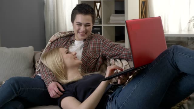 Young-blond-woman-lies-on-the-lap-of-a-brunette-and-using-red-laptop,-cosiness,-cuddles-60-fps