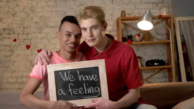 A-happy-international-gay-couple-is-sitting-on-the-couch-and-holding-a-sign.-We-have-feeling.-Look-at-the-camera.-Home-comfort-on-the-background.-60-fps