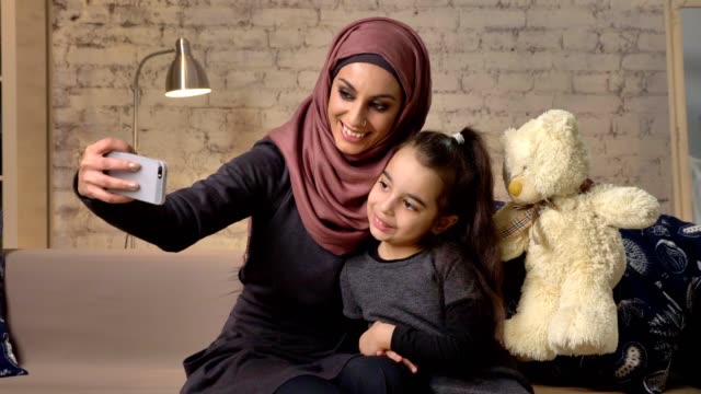 Joven-madre-hermosa-en-hijab-con-niña-en-el-sofá,-sonrientes,-aplicaciones-smartphone,-selfie-hace,-caricias,-niña-con-oso-de-peluche,-home-comfort-en-el-fondo-50-fps