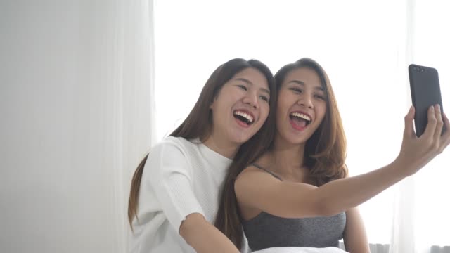 Schöne-junge-asiatische-Frauen-LGBT-lesbische-glückliche-Paar-sitzt-am-Bett-Umarmung-und-Telefon-unter-Selfie-zusammen-Schlafzimmer-zu-Hause-benutzen.-LGBT-lesbisches-Paar-gemeinsam-im-Haus-Konzept.-Schöne-Zeit-zu-Hause-verbringen.