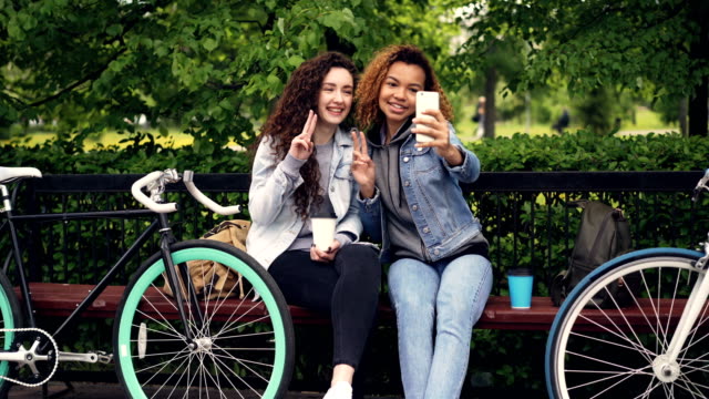 Chicas-bastante-descuidadas-están-tomando-selfie-con-smartphone-posando-y-la-celebración-para-llevar-café-sentado-en-el-parque-con-las-bicicletas.-Estilo-de-vida-moderno,-gente-alegre-y-concepto-de-tecnología.