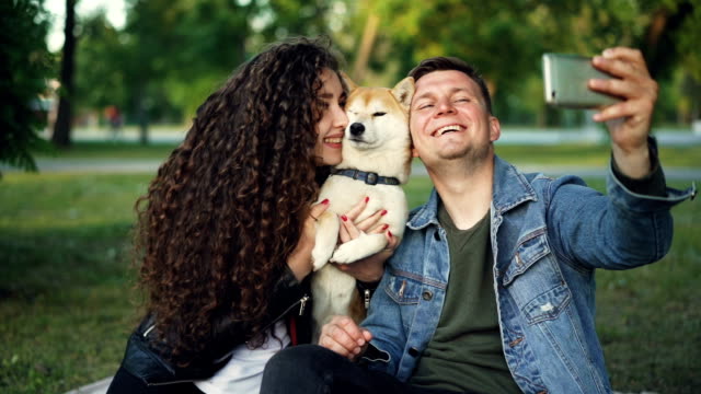 Slow-motion-of-happy-pair-girl-and-guy-taking-selfie-with-cute-dog-posing-and-kissing-animal-holding-smartphone.-Modern-technology,-parks-and-leisure-concept.