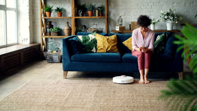 Attractive-woman-is-turning-on-robotic-vacuum-cleaner-sitting-on-sofa-then-using-smartphone-and-resting-while-device-is-cleaning-the-house.-Technology-and-apartments-concept.