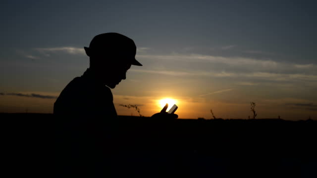 silhouette-of-a-boy-uses-a-tablet-at-sunset-in-the-field,-reads-something-on-the-tablet