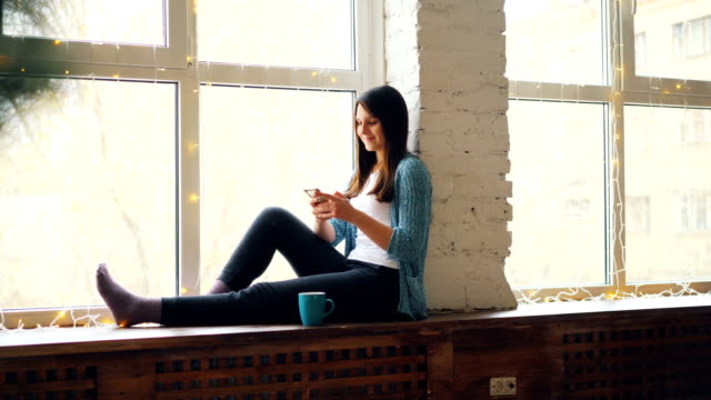 Fröhliche-junge-Frau-nutzt-Smartphone-sitzt-auf-der-Fensterbank-mit-Tasse-Tee-zu-Hause-entspannen-und-genießen-modernen-Technologie.-Menschen-und-Geräte-Konzept.