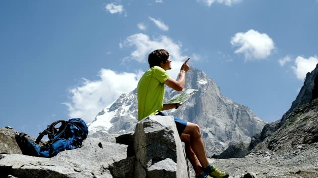 Tourist-man-hiker-looking-at-the-map-and-study-the-route-in-the-mountains-sitting-on-the-rock,-slow-motion