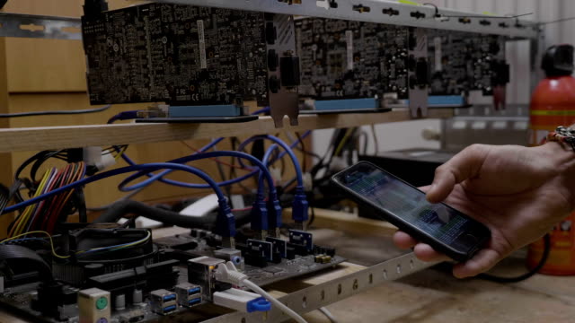 Miner-engineer-young-man-watching-mining-software-process-on-smartphone-connected-on-cryptocurrency-mining-rig