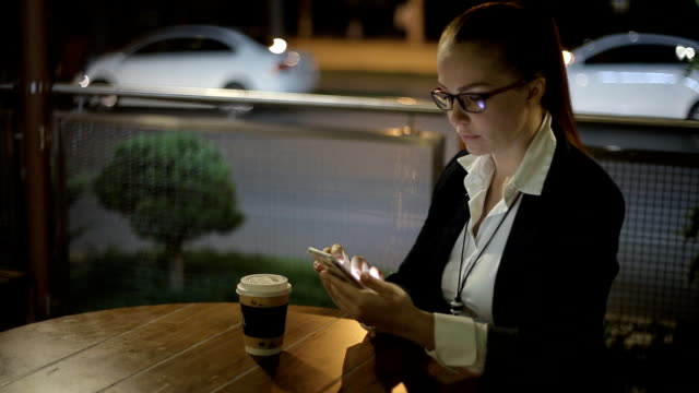 hermosa-mujer-caucásica-joven-sentada-después-del-trabajo-por-la-noche-en-un-café-en-la-calle-bebiendo-café-y-charlando-en-las-redes-sociales-usando-un-smartphone.