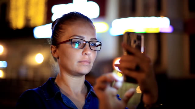 Portrait-of-young-beautiful-caucasian-woman-using-smart-phone-hand-hold-outdoor-in-the-city-night,-smiling,-face-illuminated-screenlight---social-network,-technology,-comunication-concept
