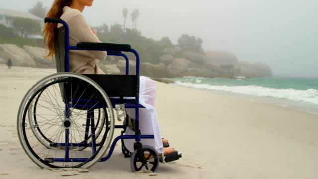 Vista-lateral-de-la-joven-mujer-caucásica-sentada-en-silla-de-ruedas-en-la-playa-4k