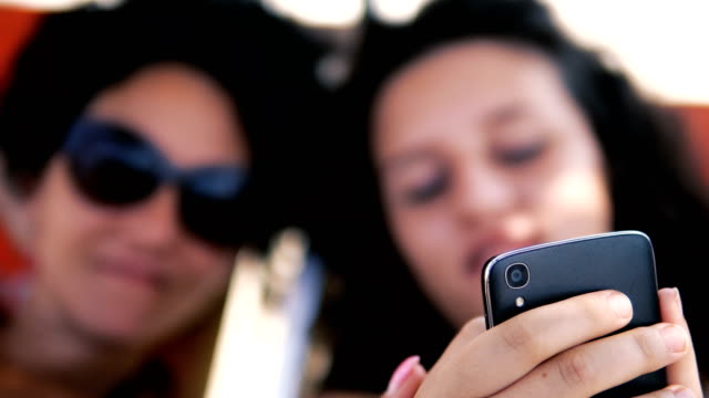 Teens-Lächeln-Vorschau-Social-Media-Beiträge-auf-Smartphone-Liegen-auf-Strandbett,-selektiver-Fokus
