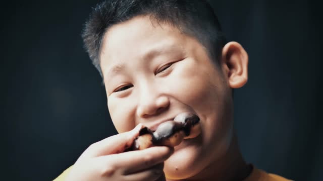 Großer-asiatischer-Vorzehn-Junge,-der-Donut-isst,-während-er-seinen-Bauch-berührt,-Zeitlupe-bei-niedrigem-Licht.