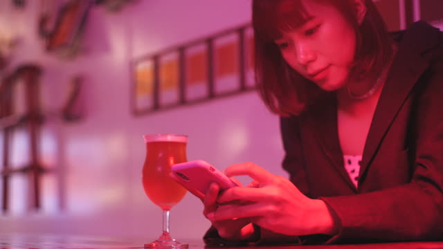 Young-asian-millennial-woman-drinking-beer-and-using-smartphone-in-nightclub-bar-at-night,-neon-pink-background