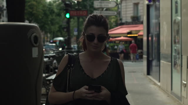 Atractiva-mujer-caucásica-con-gafas-de-sol,-pecas,-piercings-y-pelo-rojo-escribiendo-un-mensaje-de-texto-en-su-smartphone-caminando-por-la-calle,-durante-el-verano-soleado-en-París.-A-cámara-lenta.-Moda.