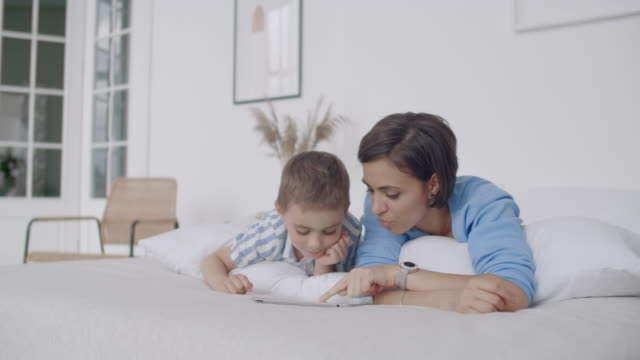 Im-weißen-Schlafzimmer-schauen-Mama-und-Sohn-auf-den-Tablet-Bildschirm-und-lachen.-Glückliche-Familie-im-Bett-am-Morgen-lesen-ein-Buch-und-klicken-Sie-auf-das-Touchpad-des-Computers