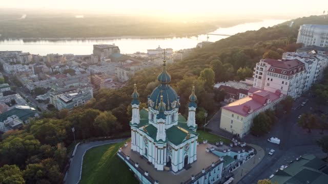 Flying-above-and-around-St.-Andrew's-Church-in-Kyiv,-Ukraine-at-sunrise.-The-church-is-now-belongs-to-Orthodox-Ecumenical-Patriarchate-of-Constantinople.-Podil-District-and-Dnieper-River-are-in-the-background.-Aerial,-4K