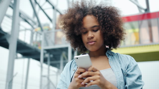 Chica-afroamericana-feliz-mirando-la-pantalla-del-teléfono-inteligente-sonriendo-al-aire-libre
