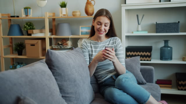 Chica-bonita-usando-el-teléfono-inteligente-moderno-tocando-la-pantalla-riendo-en-el-sofá-en-casa