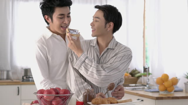 Pareja-asiática-gay-aplicando-chocolate-en-el-pan-por-la-mañana-en-casa.-Chico-gay-emoción-feliz-y-abrazar-el-aniversario-de-amor-juntos.-Concepto-de-estilo-de-vida,-familia,-gay-y-bisexual.