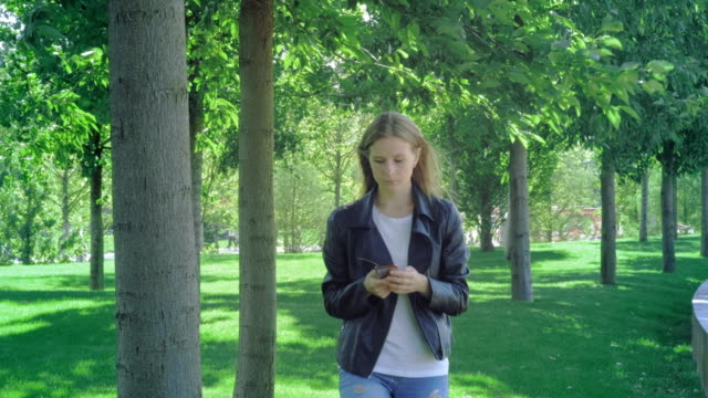 Front-view-of-a-perplexed-woman-walking-checking-smart-phone-content-and-then-stops-looking-at-camera-in-a-park.-A-young-girl-in-street-clothes-looks-at-her-smartphone-in-surprise.-Shocking-message