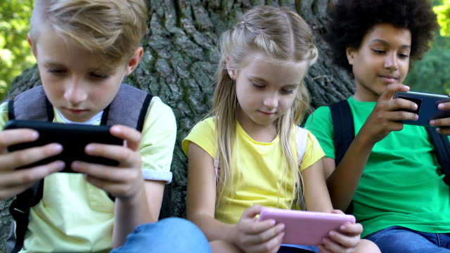Kinder-spielen-Smartphone-Spiele-sitzen-unter-Baum-im-Park,-Gadget-Sucht