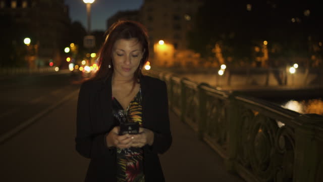 Feliz-atractivo-elegante-mujer-moderna-caucásica-usando-vestido-de-flores,-chaqueta-negra-y-pelo-rojo-caminando-por-la-calle-y-escribiendo-un-mensaje-de-texto-en-su-teléfono-inteligente-por-la-noche.-Paris-4K-UHD.