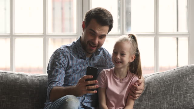 Dad-embrace-kid-daughter-having-fun-using-smartphone-on-sofa